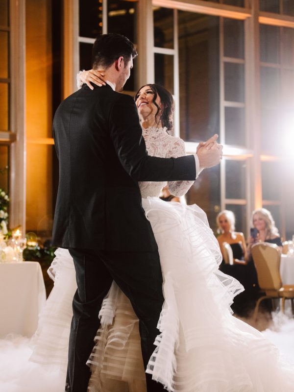 Snohomish Wedding Photographer GSquared Weddings A bride and groom share a joyful first dance at their wedding reception, smiling at each other. The bride wears a ruffled white gown and the groom is in a black suit. Guests are seated at tables in the background. Serving Seattle, Snohomish and Orlando