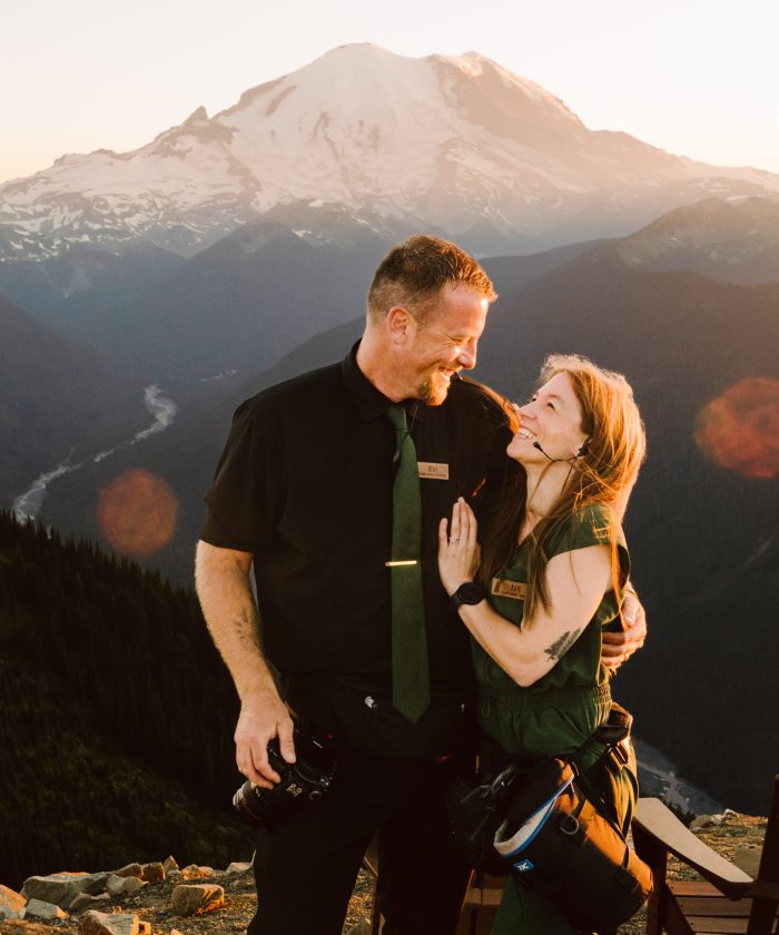 GSquared Weddings Photography A man and woman in matching dark outfits smile and embrace on a mountain overlook at sunset, perfect for an About page created with Elementor, with a snow-capped peak in the background and sunlight creating lens flares around them. GSquared Weddings | 47.9129° N, 122.0982° W | Serving Snohomish, Seattle , Orlando & Beyond