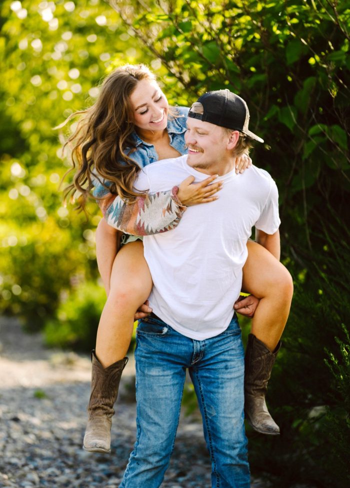 Snohomish Wedding Photographer GSquared Weddings A smiling man in a white t-shirt and jeans gives a piggyback ride to a laughing woman in boots and a denim jacket. They are outdoors on a sunny day surrounded by greenery. Serving Seattle, Snohomish and Orlando