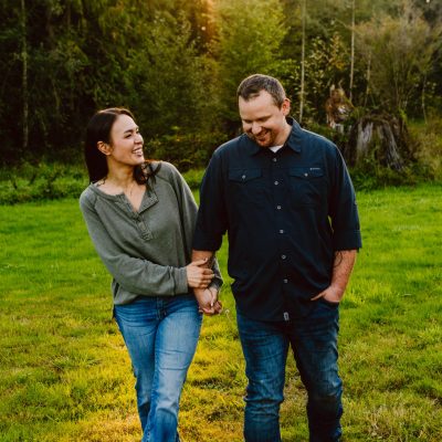 Snohomish Wedding Photographer GSquared Weddings A couple is walking hand in hand on a grassy field surrounded by trees. The woman, wearing a green sweater, smiles and looks at the man, who is wearing a dark blue shirt. Sunlight filters through the trees in the background. Serving Seattle, Snohomish and Orlando