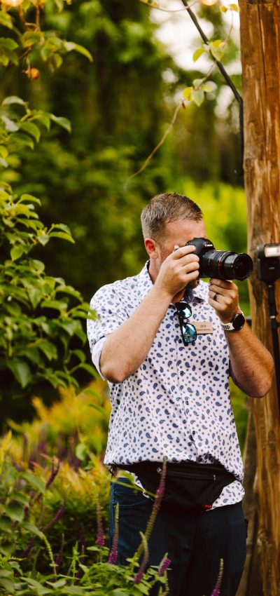 GSquared Weddings Photography A man wearing a white shirt with blue patterns is taking a photo with a camera in a lush garden, perfect for an About Elementor page. He has sunglasses hanging from his shirt and stands next to wooden posts and green foliage. GSquared Weddings | 47.9129° N, 122.0982° W | Serving Snohomish, Seattle , Orlando & Beyond