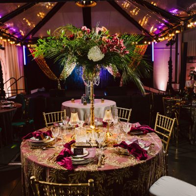 Snohomish Wedding Photographer GSquared Weddings Elegant banquet setup with a round table adorned with floral centerpiece, vibrant pink napkins, and gold chairs. Ambient lighting and clear tent canopy create a warm, luxurious atmosphere. Serving Seattle, Snohomish and Orlando
