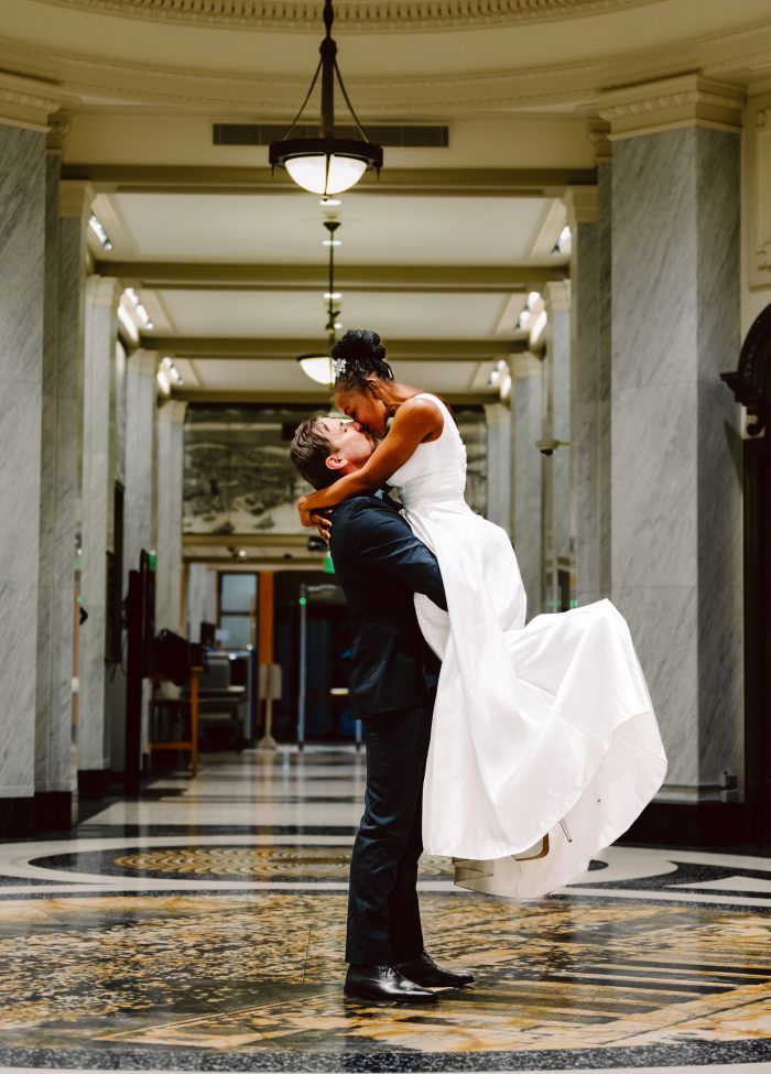 GSquared Weddings Photography A groom in a dark suit lifts and embraces his bride, who wears a white dress, in an elegant hallway with marble columns and ornate floors—capturing the joy and intimacy often found at celebrations inspired by affordable elopement prices. GSquared Weddings | 47.9129° N, 122.0982° W | Serving Snohomish, Seattle , Orlando & Beyond