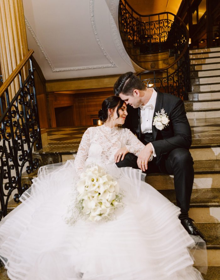 Snohomish Wedding Photographer GSquared Weddings A bride in a white lace gown and a groom in a black tuxedo sit close together on elegant stairs, holding a bouquet of white flowers. Captured by Seattle Wedding Photography, they smile affectionately as they rest their heads together. Serving Seattle, Snohomish and Orlando