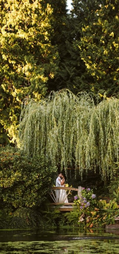 GSquared Weddings Photography A couple stands embracing on a small wooden bridge under a weeping willow, surrounded by lush greenery and flowers by the pond—just like a beautifully designed WordPress site using Elementor, with sunlight streaming through tall trees in the background. GSquared Weddings | 47.9129° N, 122.0982° W | Serving Snohomish, Seattle , Orlando & Beyond