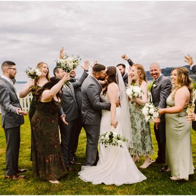 Port Gamble Summer Wedding Snohomish Wedding Photographer GSquared Weddings A bride and groom share a kiss in the center of a joyful summer wedding party. The group, dressed in suits and dresses, celebrates with raised arms and bright smiles. A white fence and the overcast sky of Port Gamble serve as the backdrop. Serving Seattle, Snohomish and Orlando
