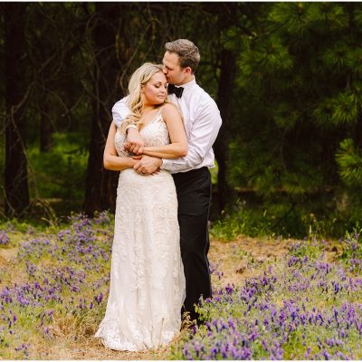 Snohomish Wedding Photographer GSquared Weddings A couple stands in a field of purple flowers near a quaint winery, surrounded by trees. The man, in a white shirt and black pants, embraces the woman, who wears a white lace dress. They both appear serene and content. Serving Seattle, Snohomish and Orlando
