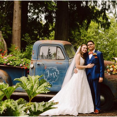 Stanwood Backyard Wedding Snohomish Wedding Photographer GSquared Weddings In a charming Stanwood setting, a bride in a white dress and a groom in a blue suit stand happily in front of a vintage blue pickup truck adorned with flowers. The truck is parked amid the lush beauty of their backyard garden, creating the perfect backdrop for their wedding day. Serving Seattle, Snohomish and Orlando